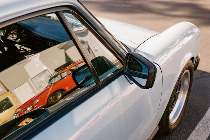 A white car is parked with a reflection of an orange car visible in its window.