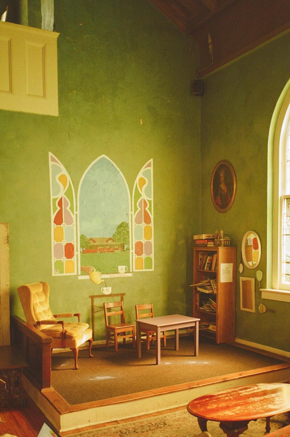 A room with green walls features a painted mural of a window, a small table with chairs, a bookshelf, and an armchair.