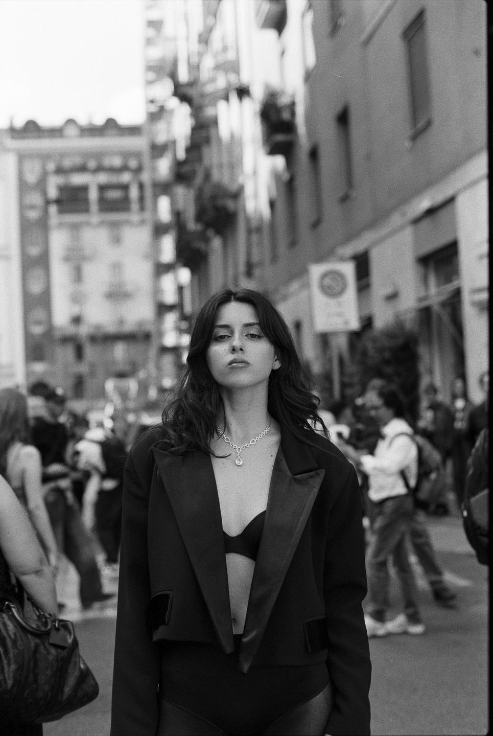 A woman wearing a blazer and bra stands on a city street with buildings and people in the background.