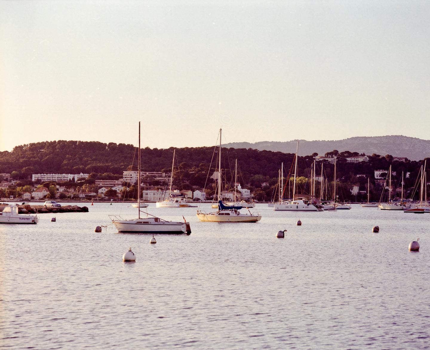 Numerous sailboats are moored in a calm bay with a town and forested hills in the background during sunset.