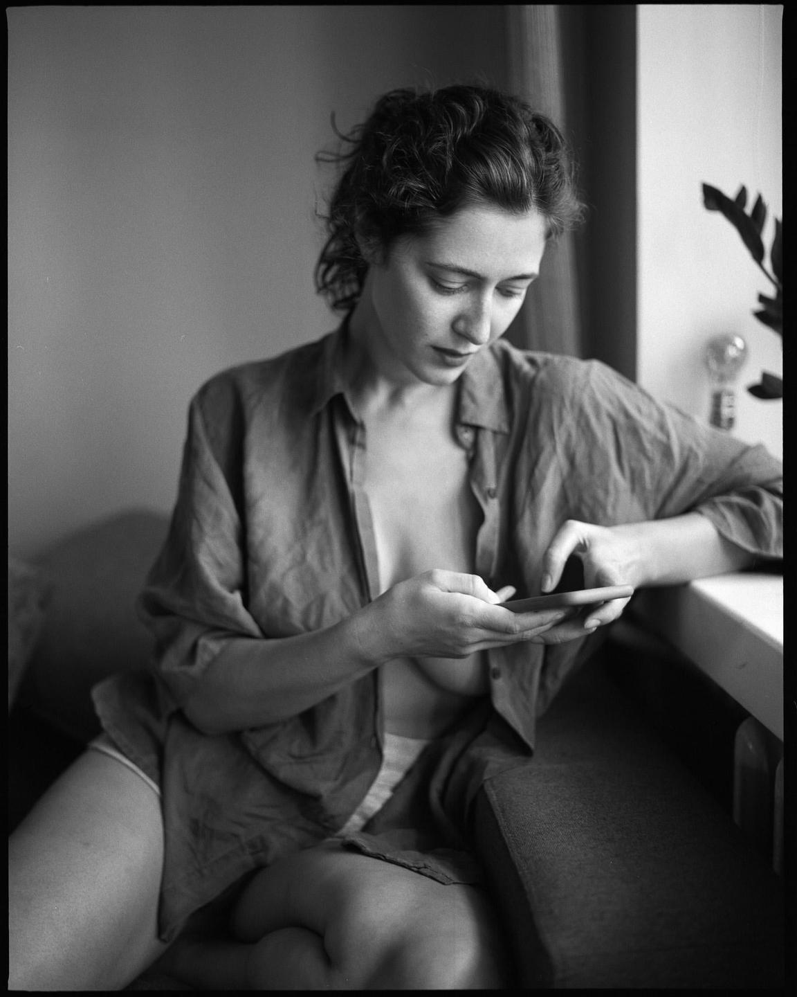 A woman in an unbuttoned shirt sits and looks at her phone.