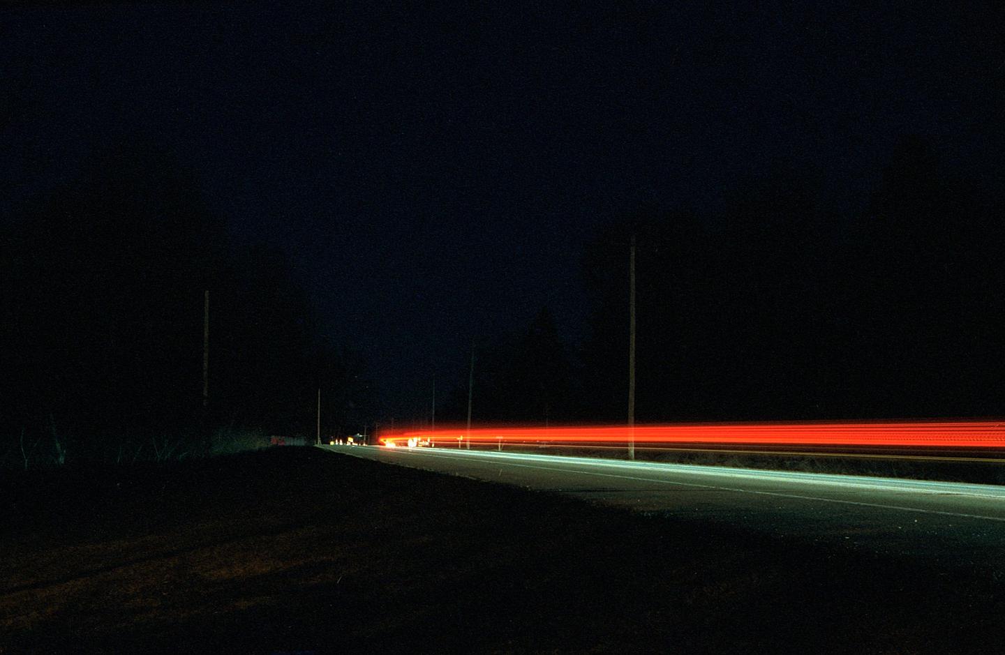 Red and white light trails from vehicles illuminate a dark road at night.