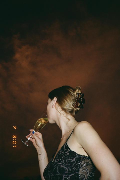 A woman in a dark dress drinks from a champagne flute against a dark, cloudy sky.