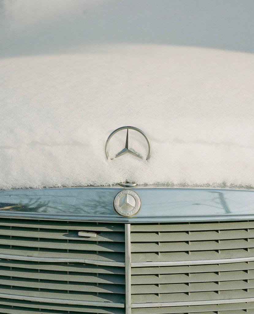 A close-up shot shows the front of a car covered in snow, with the Mercedes-Benz emblem visible.
