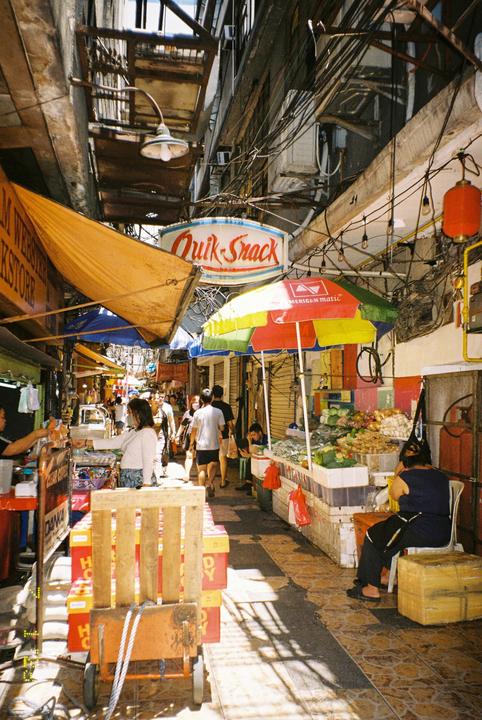 A narrow street market is bustling with people, vendors, and colorful stalls under a bright sky.
