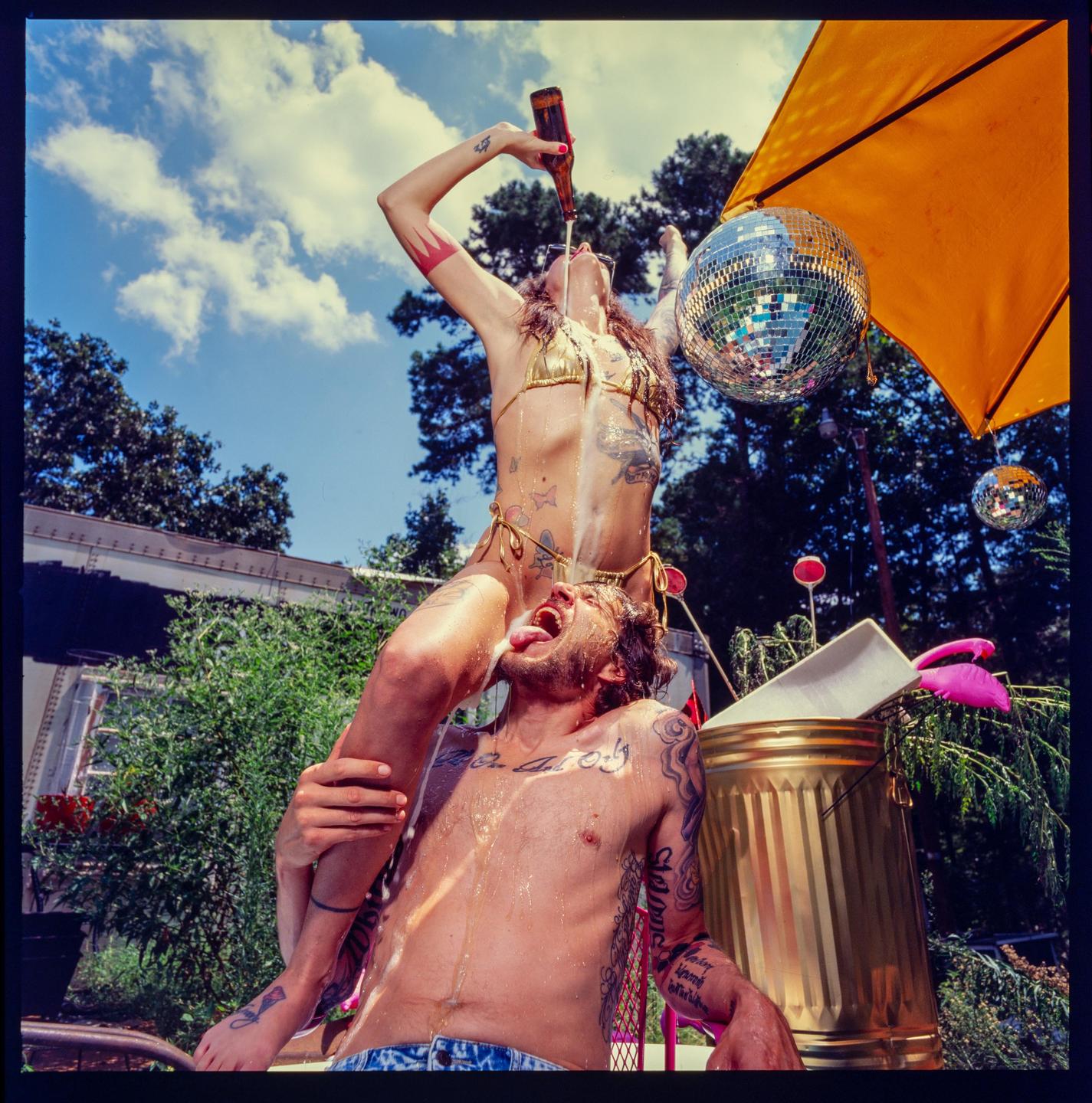 A woman in a gold bikini pours beer into a man's mouth while sitting on his shoulders outdoors.