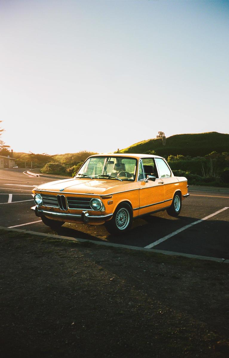 A classic orange car is parked on an asphalt lot with a grassy hill and bright sky in the background.
