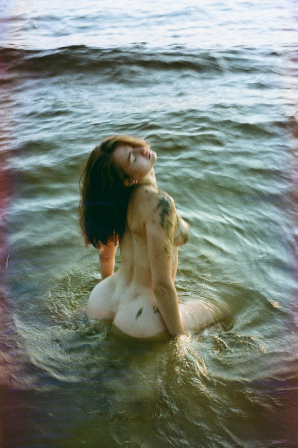 A woman with her back to the viewer sits in the water with her head tilted back and eyes closed.