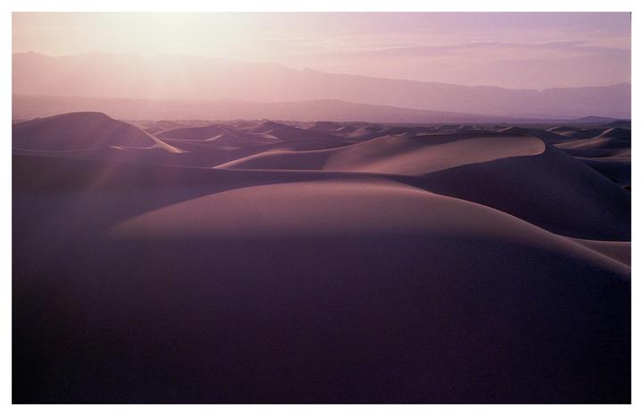 The sun shines over a vast desert landscape with rolling sand dunes and distant mountains.