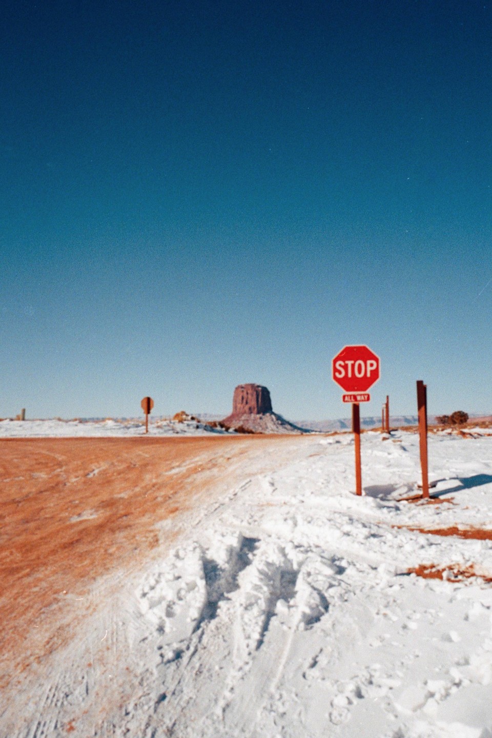 A stop sign stands in a snowy landscape with a red dirt road and a large rock formation in the distance under a clear blue sky.
