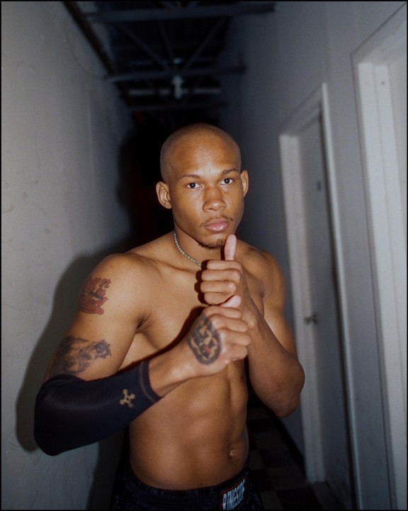 A man with a shaved head and tattoos poses with his fists together in a dimly lit hallway.