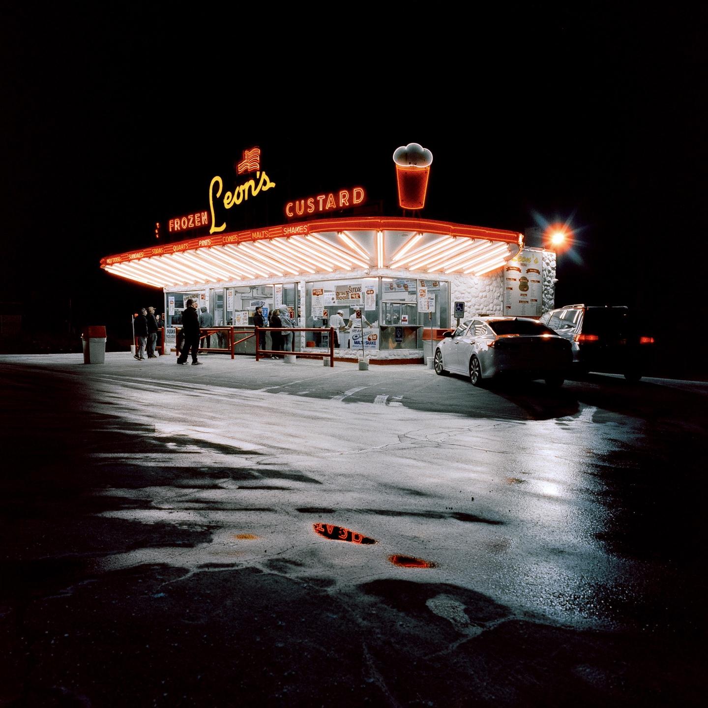 A brightly lit custard stand with neon signs stands out against the dark night, with cars and people gathered around.
