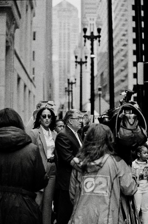 A crowd of people, including a woman in sunglasses and a man in glasses, are gathered on a city street with tall buildings and streetlights in the background.