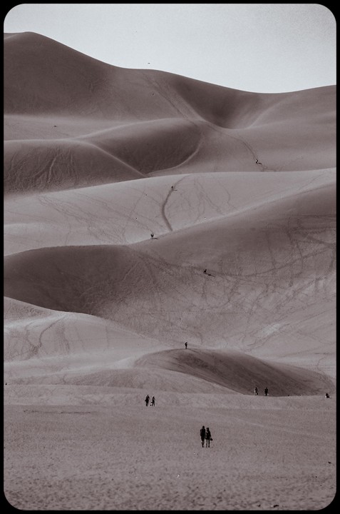 Several people walk on large sand dunes with many footprints and trails.