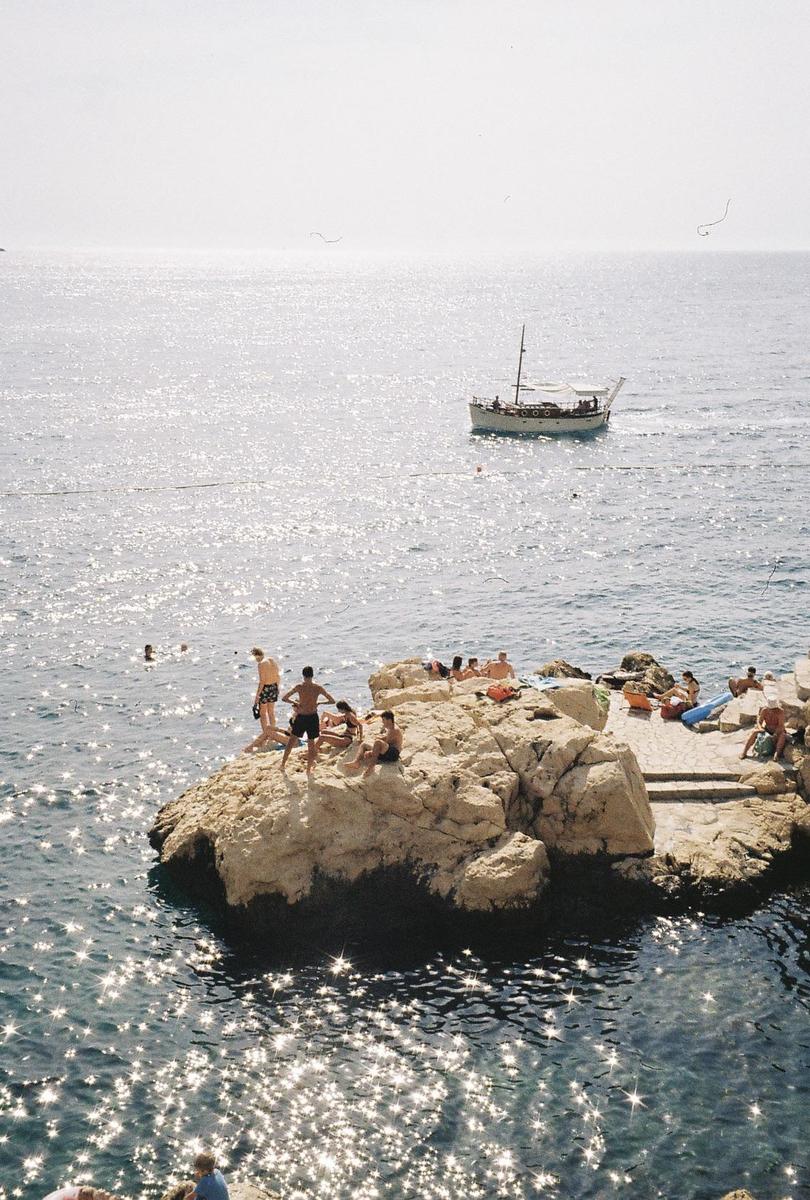 People are swimming and sunbathing on rocks in the Adriatic Sea, with a boat in the distance.