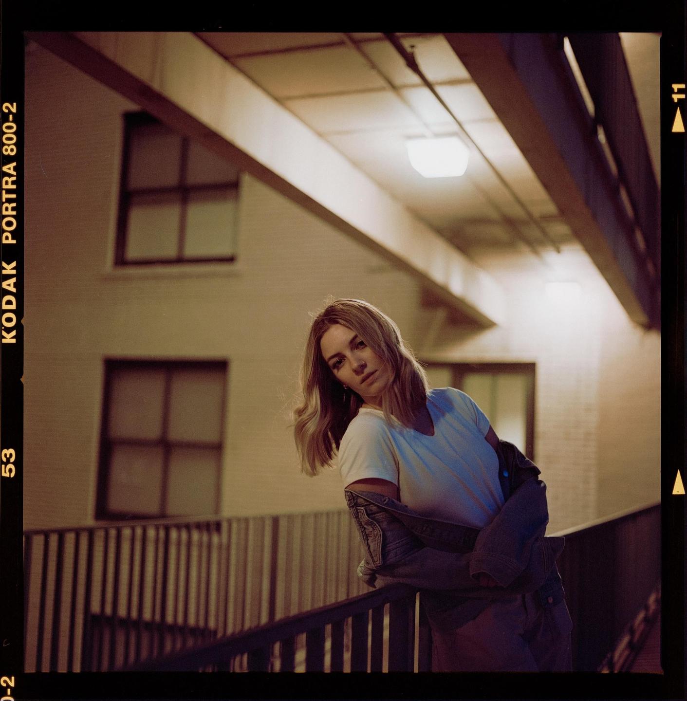A woman leans on a railing at night with a building and overhead lights in the background.
