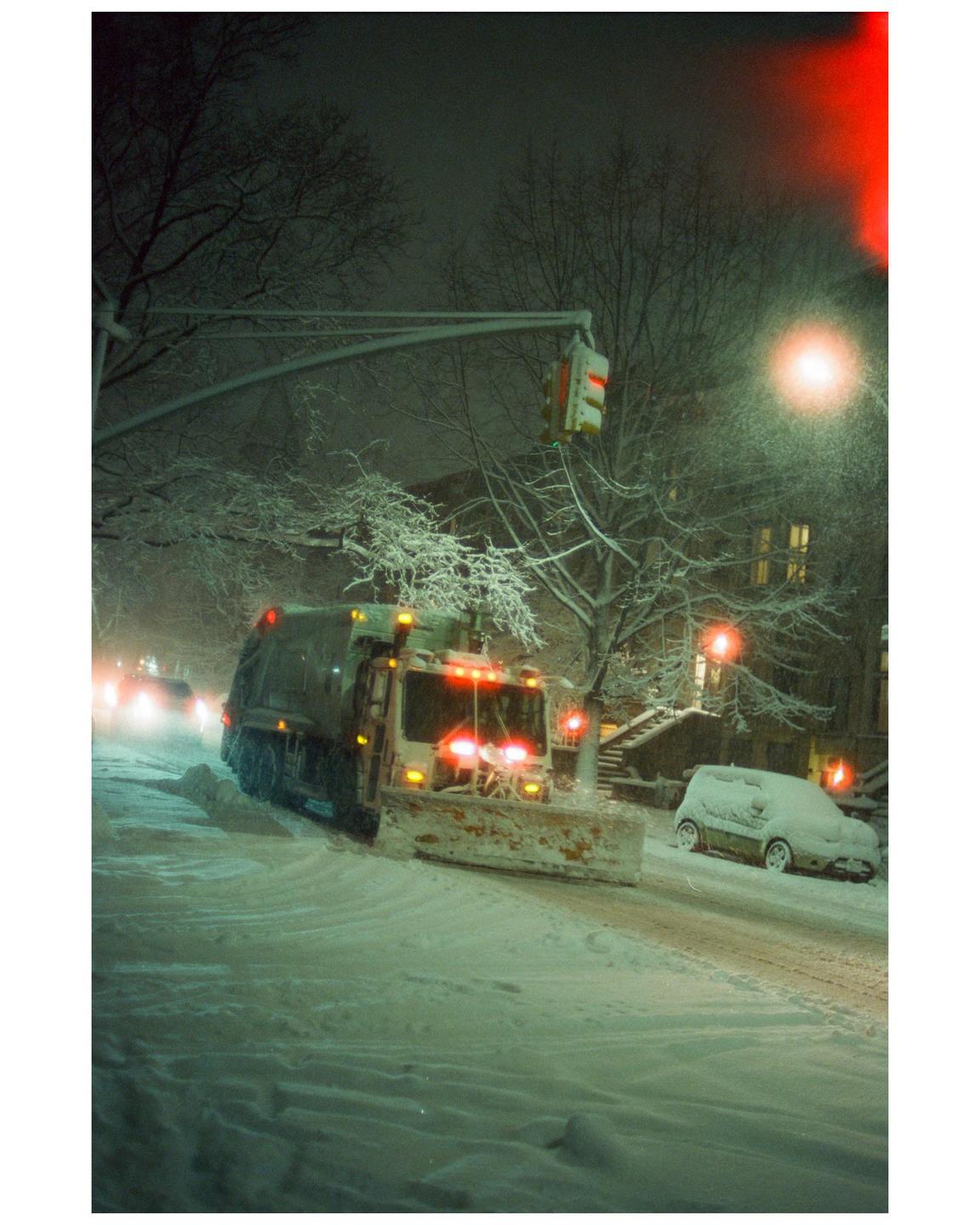 A snowplow clears a snow-covered street at night, with a car parked on the side of the road.