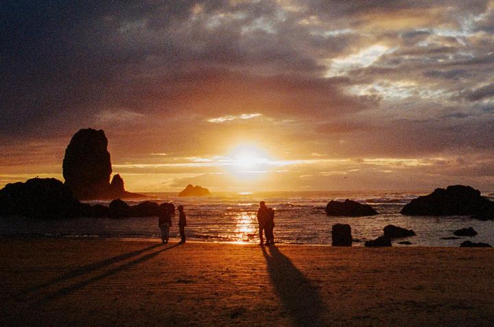 A group of people stand on a beach at sunset, with large rock formations in the ocean and dramatic clouds in the sky.