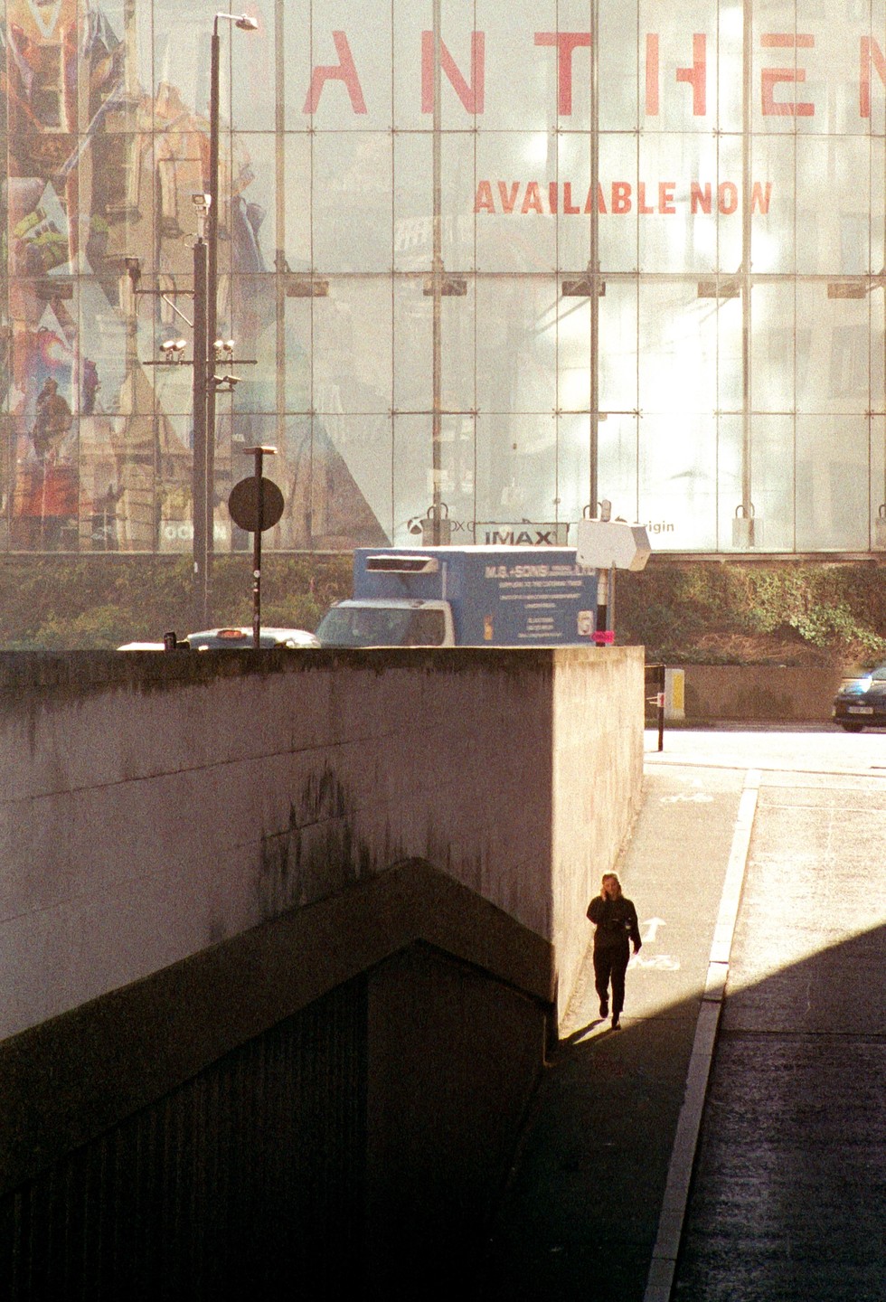 A woman walks along a sunlit path next to a concrete wall, with a large glass building reflecting the surroundings in the background.