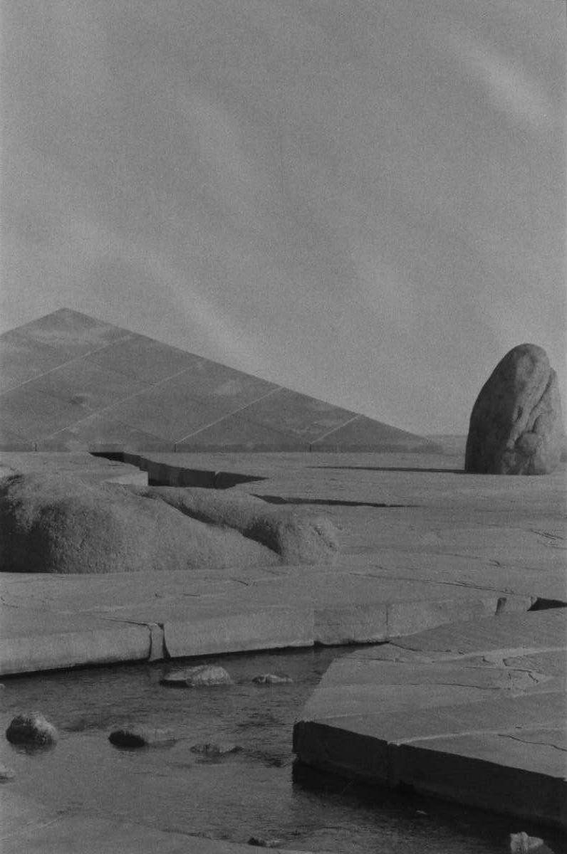 A stream flows through a rocky landscape with a large pyramid-like structure and a boulder under a bright sky.