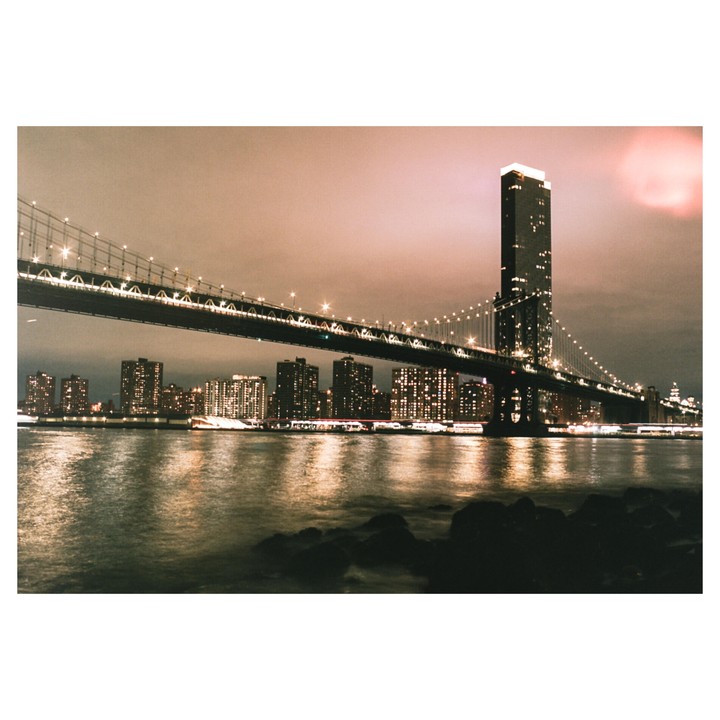 A suspension bridge with lights crosses a body of water with a city skyline and a tall building in the background at night.