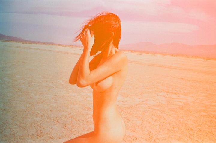 A nude woman with her hands near her face is kneeling in a desert landscape with mountains in the distance.