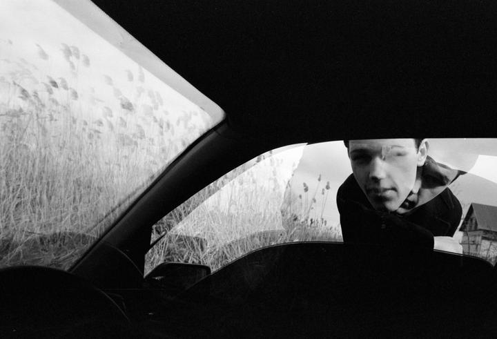 A man leans into a car window, with tall grass and a building visible in the background.