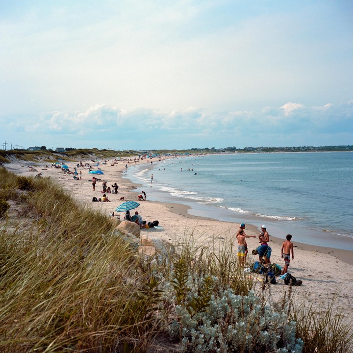 A wide sandy beach is filled with people swimming, sunbathing, and playing under a partly cloudy sky.