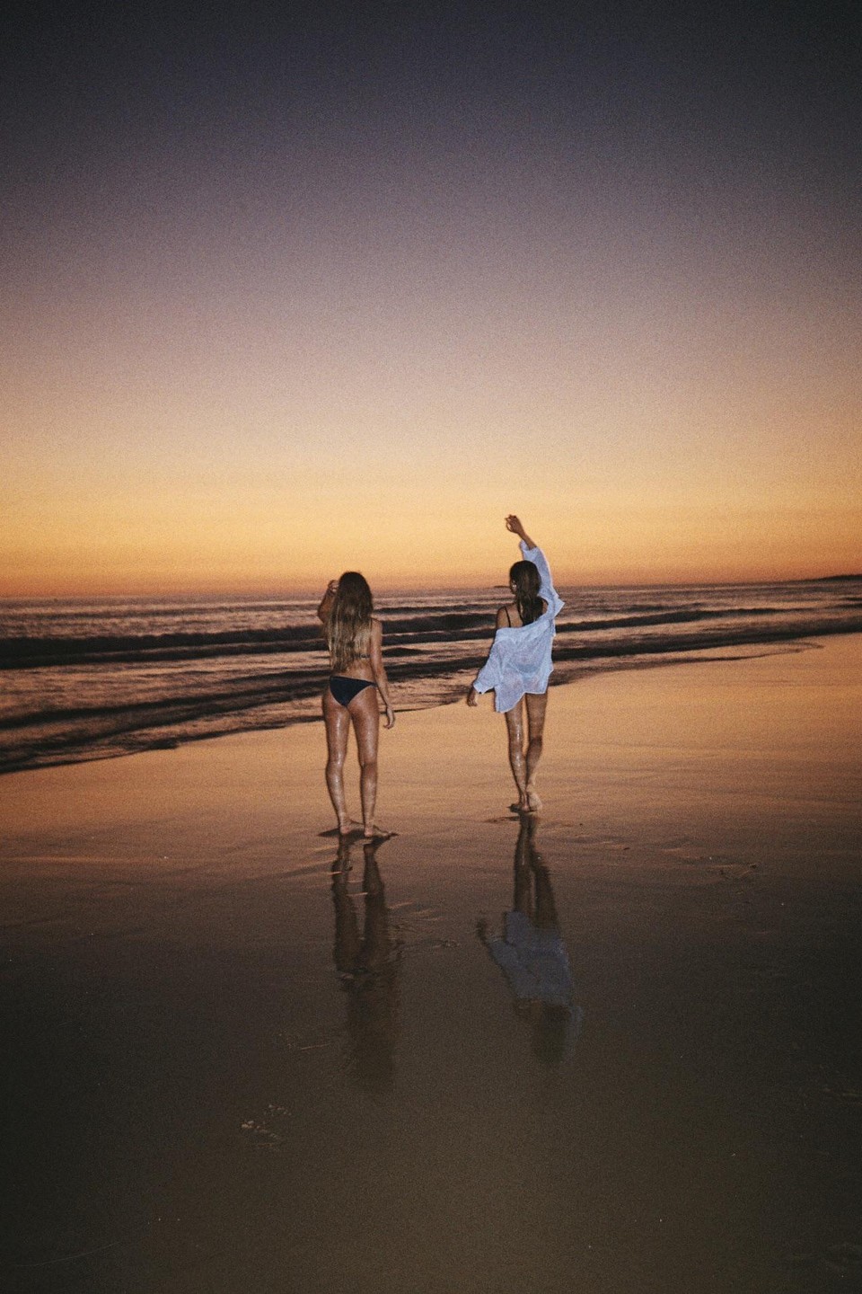 Two women stand on a beach at sunset, with their reflections visible in the wet sand.