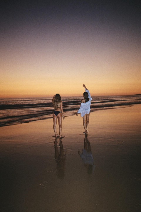 Two women stand on a beach at sunset, with their reflections visible in the wet sand.
