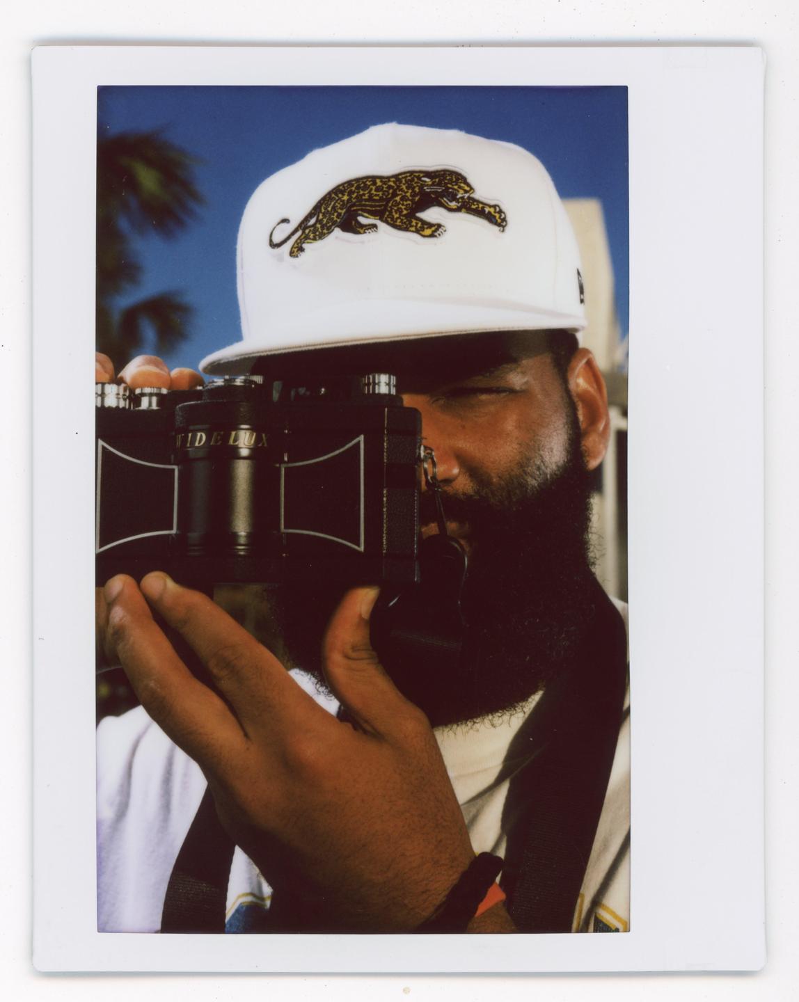 A man with a beard and a white cap with a leopard on it holds a camera up to his eye.