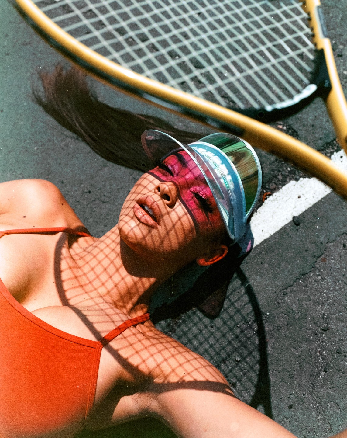 A woman with pink eye makeup and a clear visor lies on the ground with the shadow of a tennis racket over her face and body.