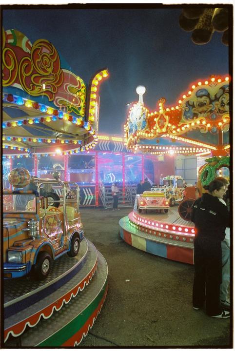 A colorful fairground ride with bright lights is captured at night, with people standing near the attractions.