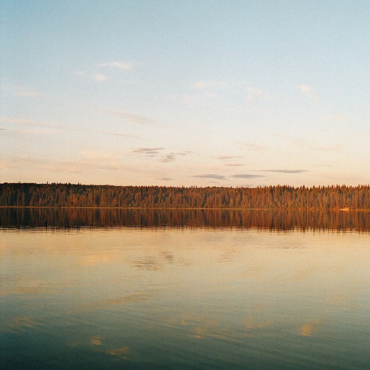 A forest reflects on the still surface of a lake under a soft, colorful sky.