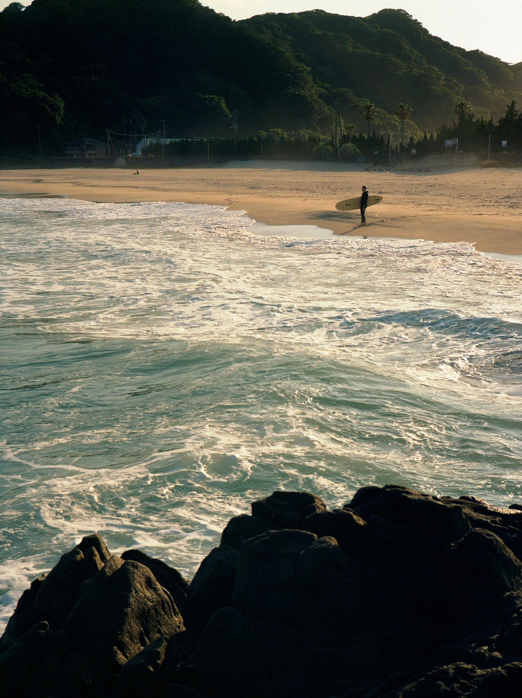 A surfer stands on a sandy beach with a surfboard, looking out at the ocean waves.