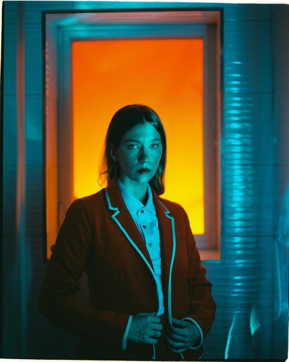 A woman in a red jacket stands in front of a window with an orange glow.