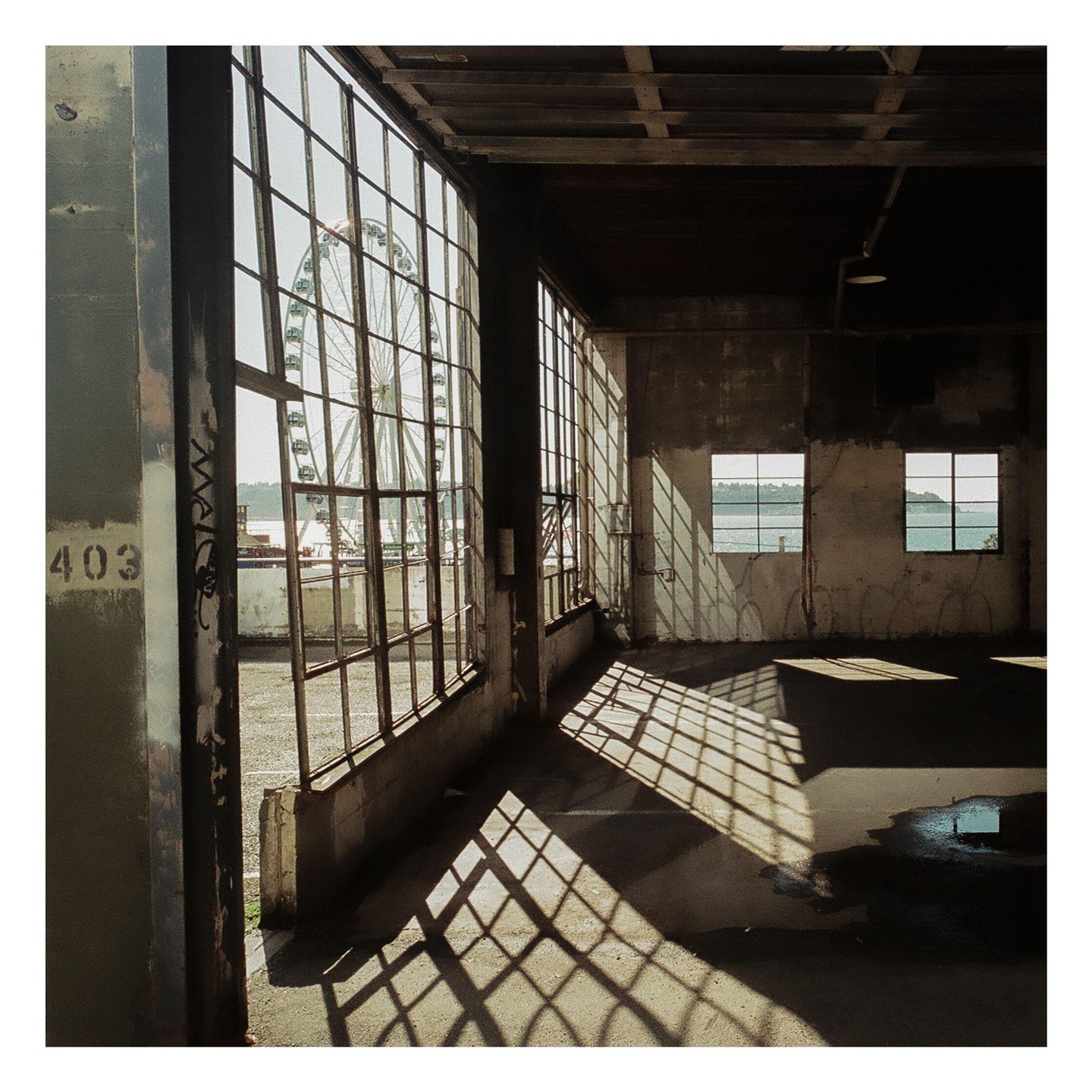 Sunlight streams through large industrial windows, casting grid shadows on the floor of an empty room, with a ferris wheel visible outside.