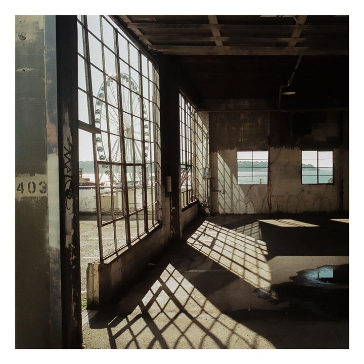 Sunlight streams through large industrial windows, casting grid shadows on the floor of an empty room, with a ferris wheel visible outside.