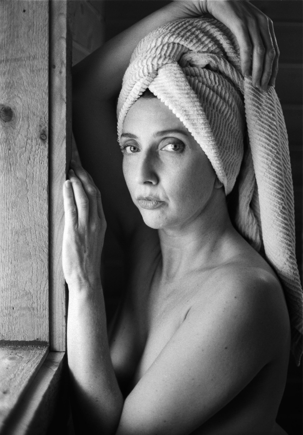 A woman with a towel wrapped around her head looks directly at the viewer, leaning against a wooden frame.