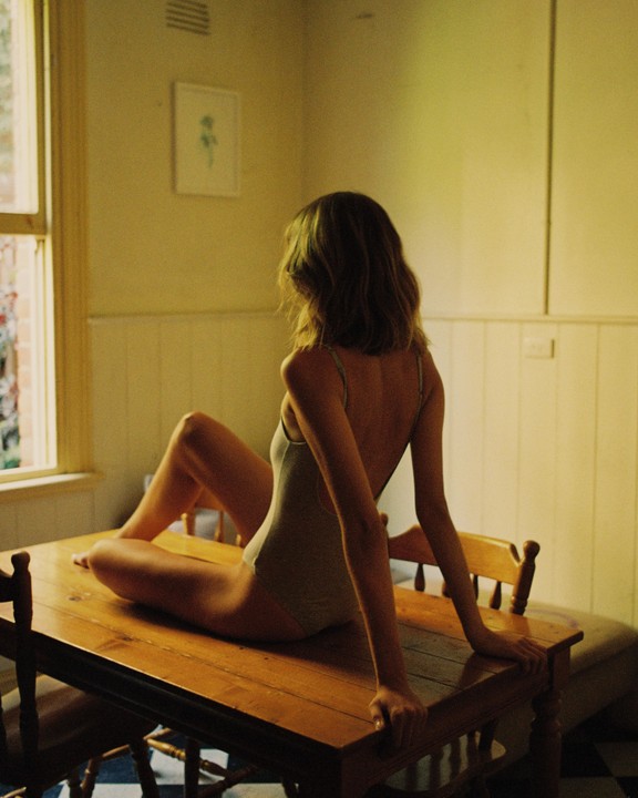 A woman in a leotard sits on a wooden table, looking out a window.