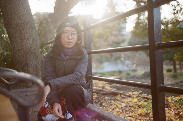 A woman wearing a beanie and glasses crouches on a concrete ledge next to a metal fence, with autumn leaves scattered on the ground and sunlight shining through trees in the background.