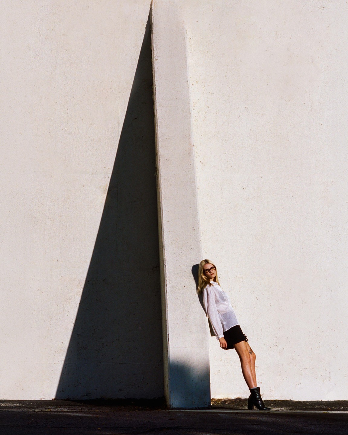 A woman in a white shirt and black skirt leans against a white wall with a large triangular shadow.