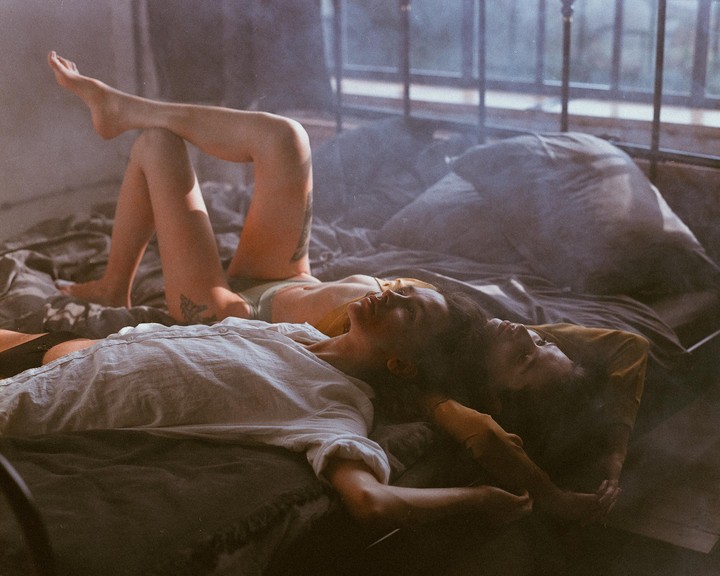 Two women are lying on a bed with light streaming in from a window.