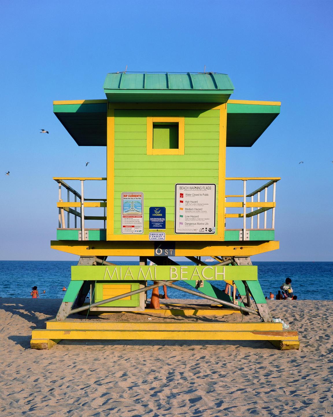 A green and yellow lifeguard stand sits on a sandy beach with the ocean and a clear blue sky in the background.