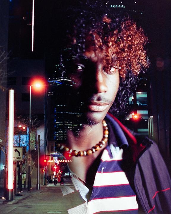 A man with curly hair and a beaded necklace is double exposed over a night street scene with buildings and traffic lights.