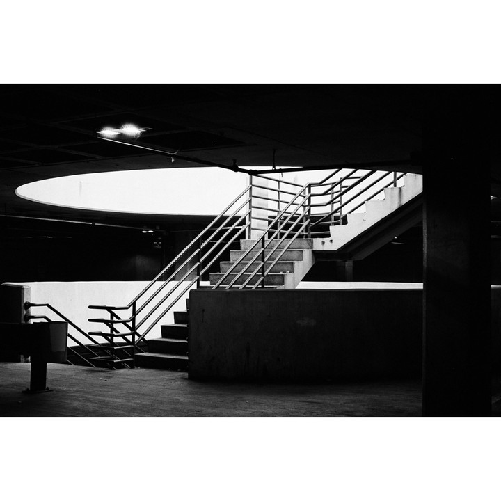 A concrete staircase with metal railings leads up from a dark parking garage toward a bright, circular opening in the ceiling.