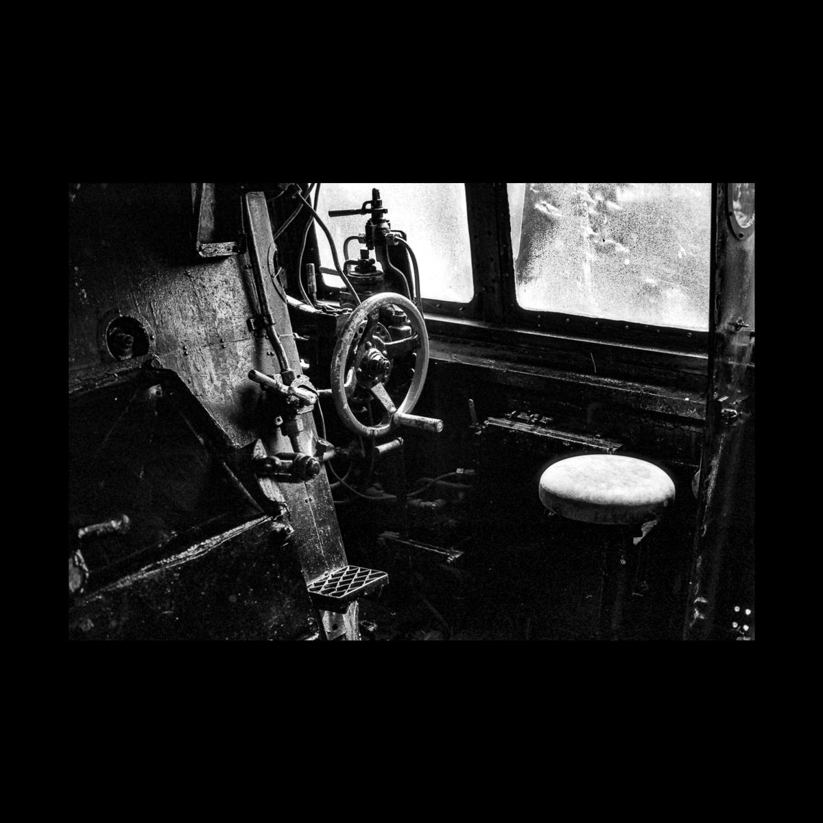 The interior of a train engine shows a steering wheel, levers, and a stool next to a window.