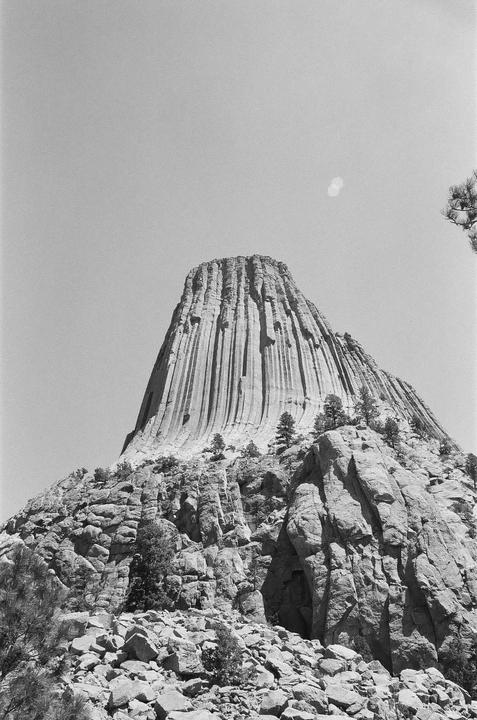 A black and white landscape shows Devils Tower rising above a rocky base with scattered trees.
