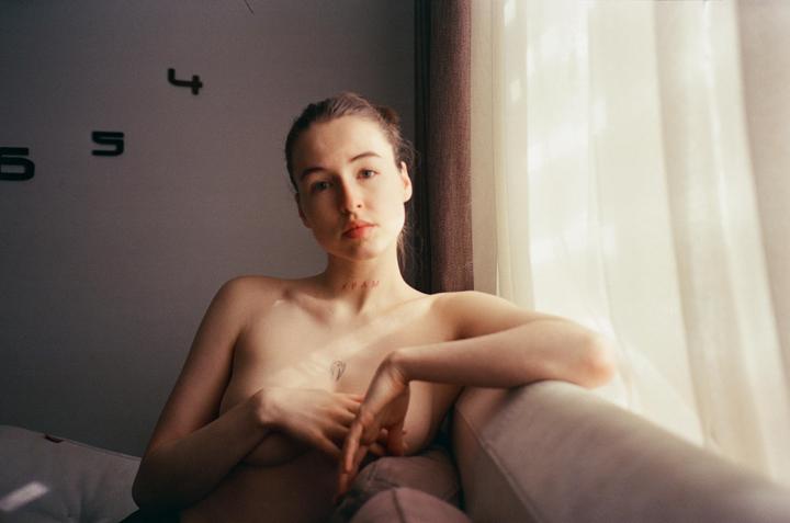 A woman with a tattoo on her chest is sitting on a couch next to a window with sheer curtains.