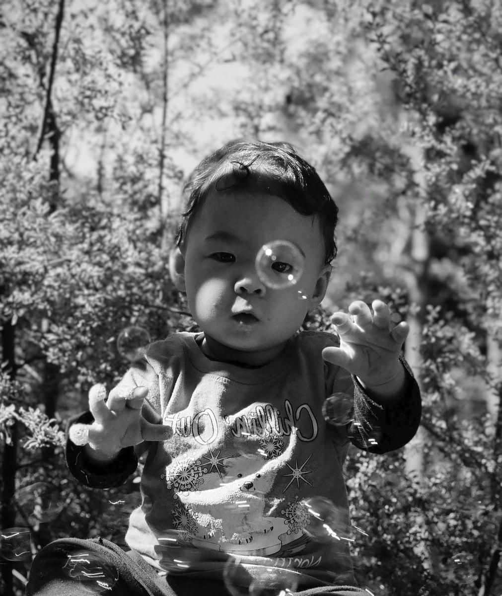 A child with dark hair and a patterned shirt reaches out towards bubbles in a sunlit outdoor setting.